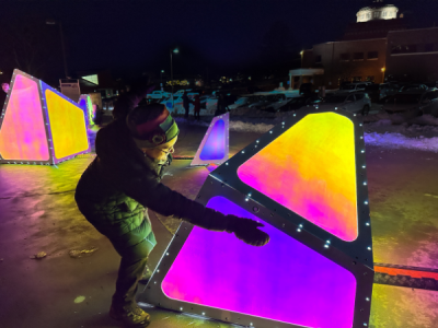 Photo of a woman in winter wear reaching out towards a large geomentric structure illuminated with a range of colors. The setting is at nightitme and other large parts of the sculptures are illuminated in the background with other visitors around them.
