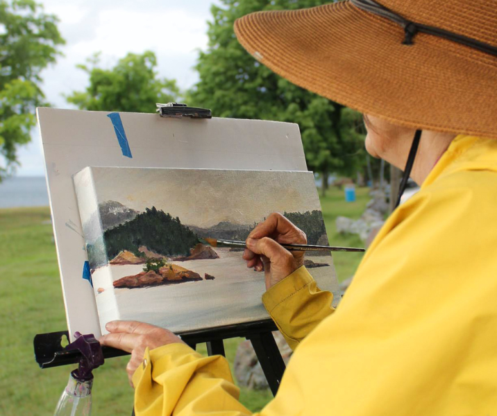 Woman in yellow jacket painting on a small canvas outside. She is standing in a grassy area overlooking Lake Superior and her painting is a panoramic view of the islands and hills of the shoreline across the bay