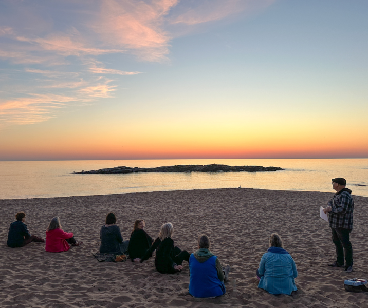 Small group of people sitting on a beach at sunrise with one person standing and looking out over the water.